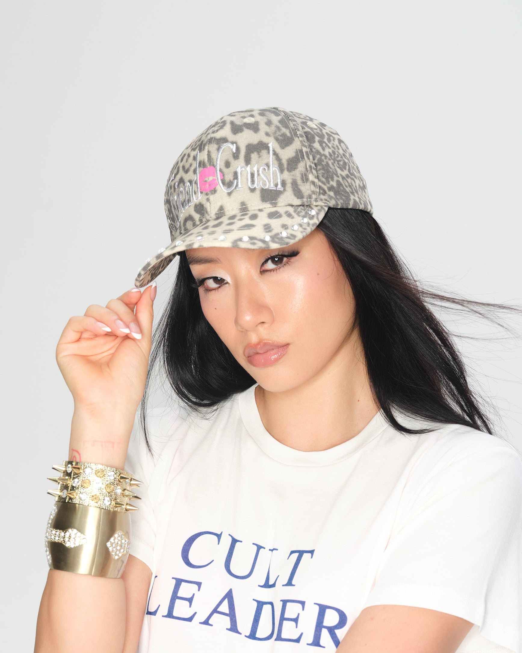 A woman wearing a leopard-print baseball cap with a pink 'Cult Leader' logo and a white graphic t-shirt, posing against a light gray background.