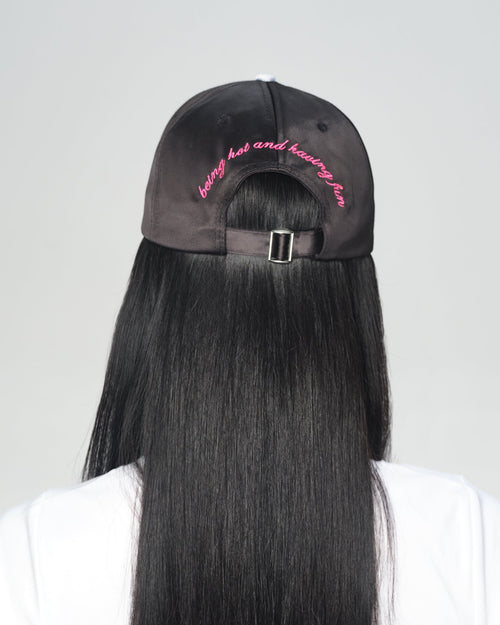 A person shown from the back wearing a black baseball cap and a white T-shirt, with very long straight black hair hanging down the back. The cap has pink 'being hot and having fun' embroidered text on the back.