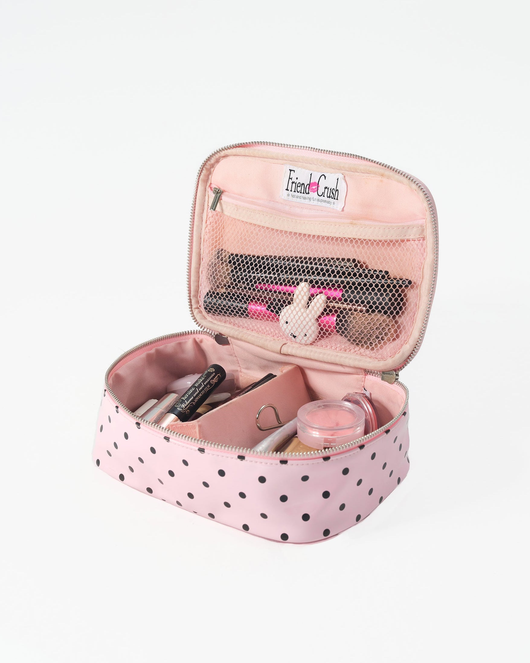 A pink zippered cosmetic bag with black polka dots, shown open to reveal its interior compartments filled with makeup items. The bag has a white 'Friend Crush' brand label on the mesh pocket.