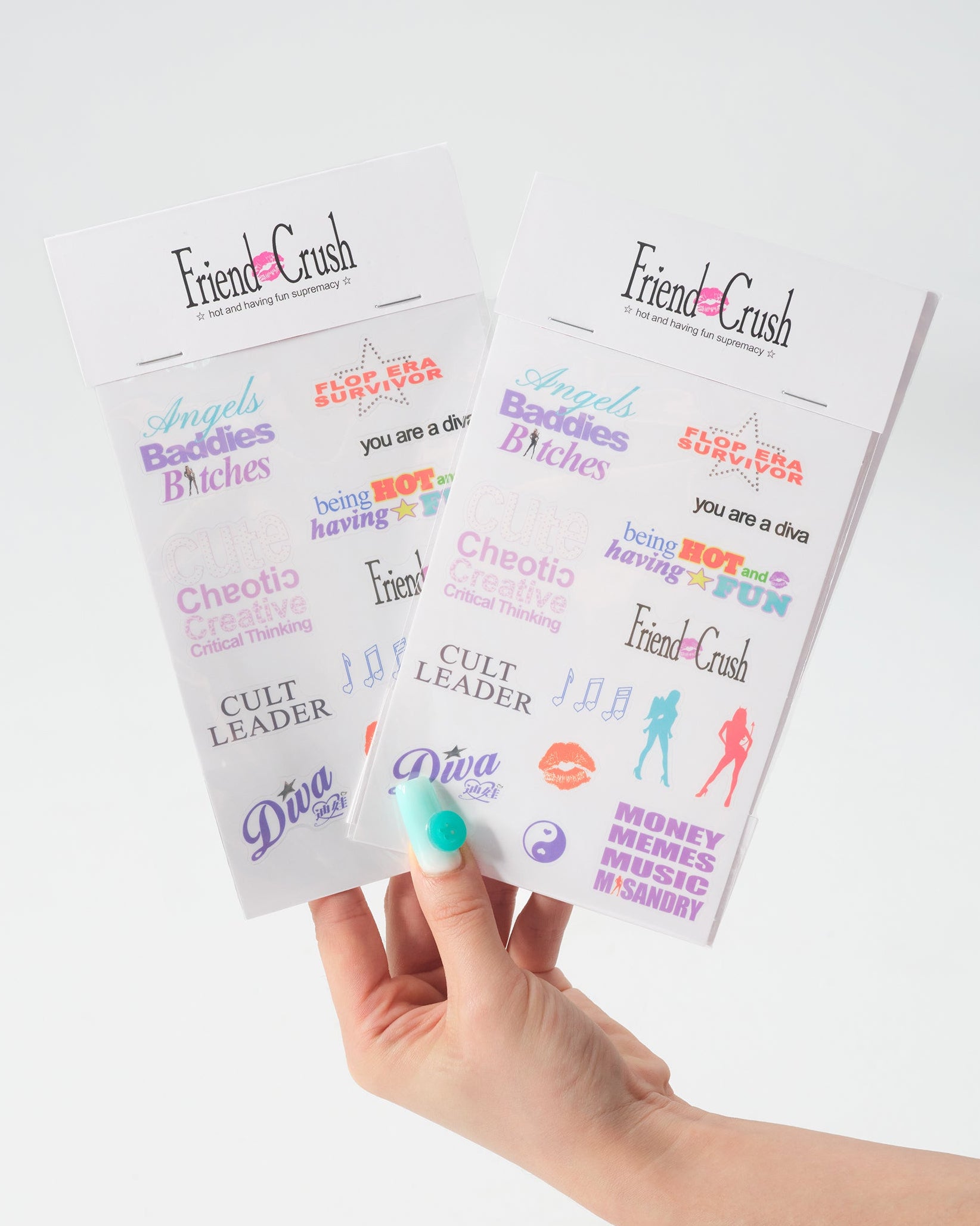 A hand with teal nail polish holds two printed sticker sheets from Friend Crush against a clean white background. The sheets feature various colorful slang and pop-culture phrases like 'Angels, Baddies & Bitches', 'Cute, Chaotic & Critical Thinking', 'Friend Crush', and 'Cult Leader'.