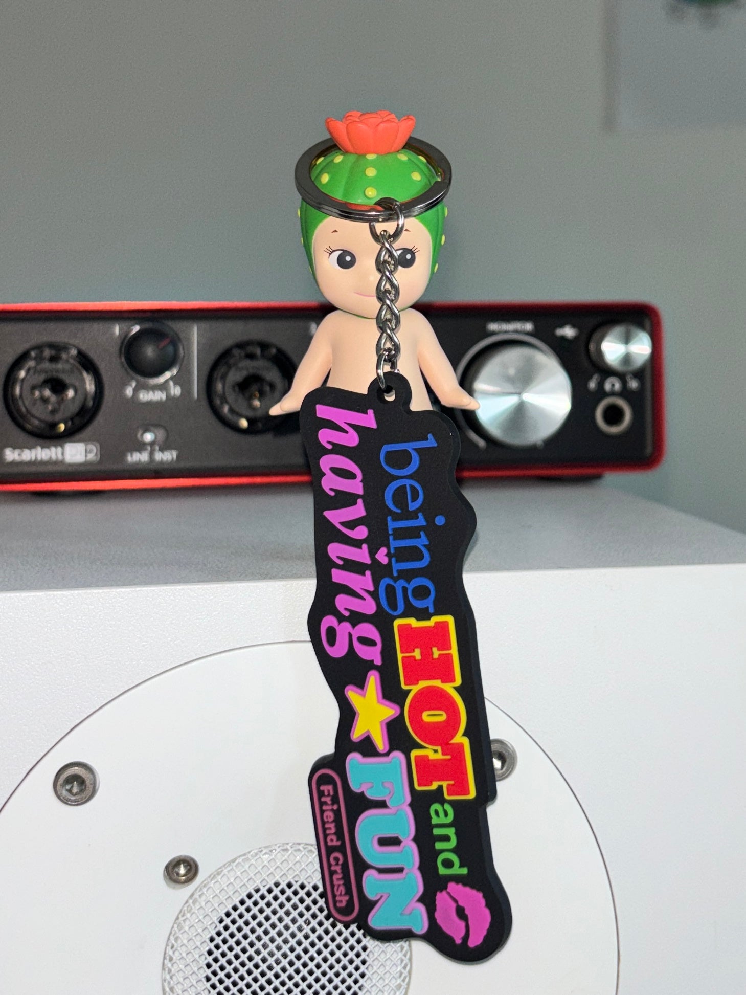 A custom rubber keychain featuring a small doll with green hair and a pink flower, attached to a black 'being HOT and FUN' tag with colorful text and graphics, set against a background with audio equipment.