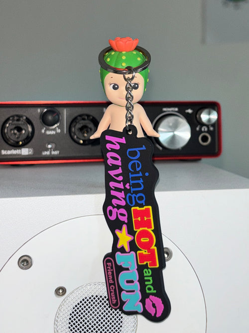A custom rubber keychain featuring a small doll with green hair and a pink flower, attached to a black 'being HOT and FUN' tag with colorful text and graphics, set against a background with audio equipment.
